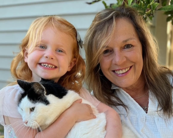 Woman and young girl holding a cat, standing outdoors with greenery and a building in the background.